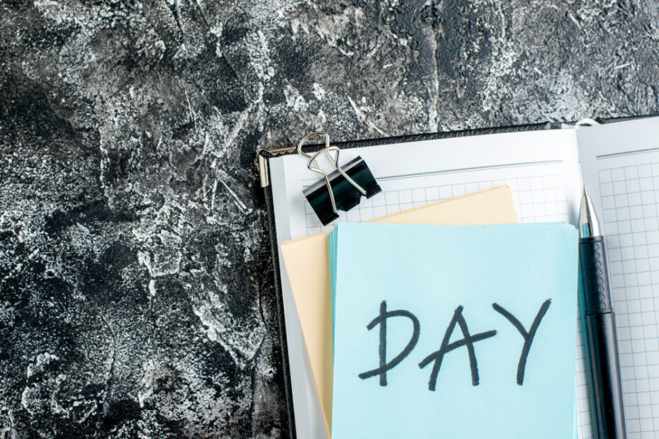 A notebook with a pen, binder clip, and a blue sticky note on top with the word "DAY" written on it, placed on a textured dark surface.