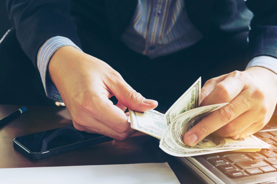 A person counting US dollar bills over a desk, with a smartphone and a calculator nearby, highlighting a financial transaction or salary calculation.