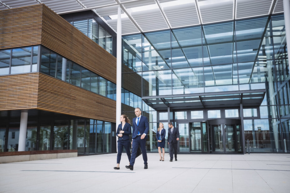 A group of business professionals walking outside a modern office building with large glass windows and sleek, wooden exterior panels. The building's open architecture and clean design reflect a contemporary corporate environment, while the people appear to be in formal attire, engaged in conversation, implying a business meeting or discussion as they approach the entrance.