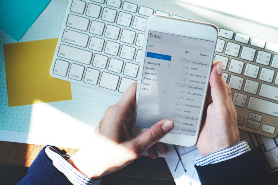 The image shows a person holding a smartphone displaying an email inbox. The individual appears to be checking or composing emails, highlighting a professional setting. The person is dressed in business attire, with a keyboard and documents visible on the desk, suggesting an office environment. The email interface on the phone shows various folders like "Inbox," "Drafts," and others, emphasizing the process of managing and organizing emails, which is essential for tasks such as sending PTO requests or handling professional communication efficiently.