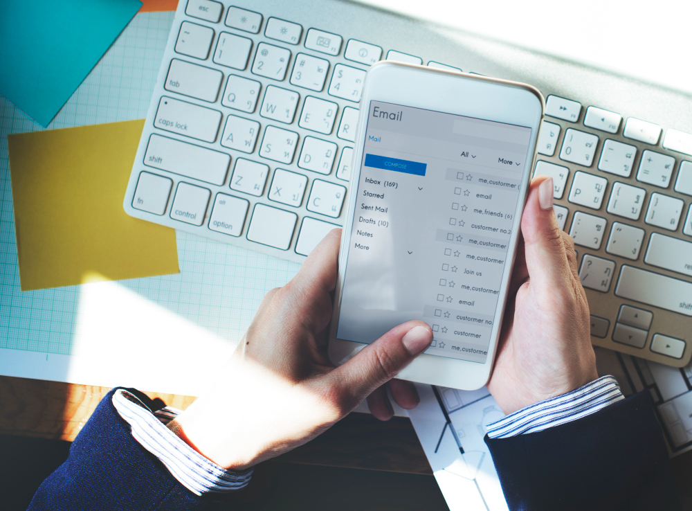 The image shows a person holding a smartphone displaying an email inbox. The individual appears to be checking or composing emails, highlighting a professional setting. The person is dressed in business attire, with a keyboard and documents visible on the desk, suggesting an office environment. The email interface on the phone shows various folders like "Inbox," "Drafts," and others, emphasizing the process of managing and organizing emails, which is essential for tasks such as sending PTO requests or handling professional communication efficiently.
