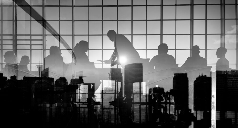 A black and white image of a group of people in a modern office, with large windows overlooking a city skyline. The scene is captured with reflections of the city superimposed on the silhouettes of the individuals, creating a layered, abstract effect. The group appears to be engaged in a meeting or discussion, with one person standing and addressing the others as sunlight streams in from the background.