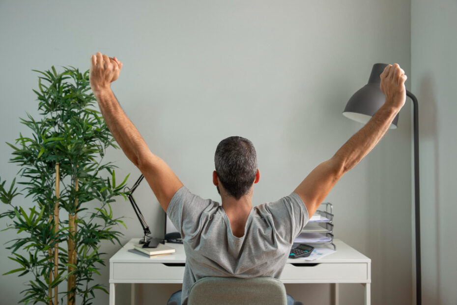 The image shows a person sitting at a desk with their arms raised triumphantly, as if stretching or celebrating. The individual, seen from behind, is wearing a gray t-shirt and appears to be taking a break in a home or office environment. The desk is equipped with a laptop, a desk lamp, a stack of folders, and some notebooks, indicating a work or study setting. To the left of the person, there is a potted plant, adding a touch of greenery to the space. The overall atmosphere suggests a moment of relief or satisfaction, possibly related to work or personal achievement.