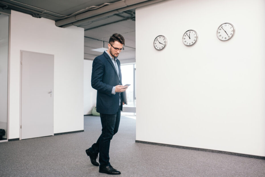 A man in a business suit walking down a hallway while looking at his smartphone. The wall to his right displays three clocks showing different times, suggesting a professional environment with a global or time-conscious focus.