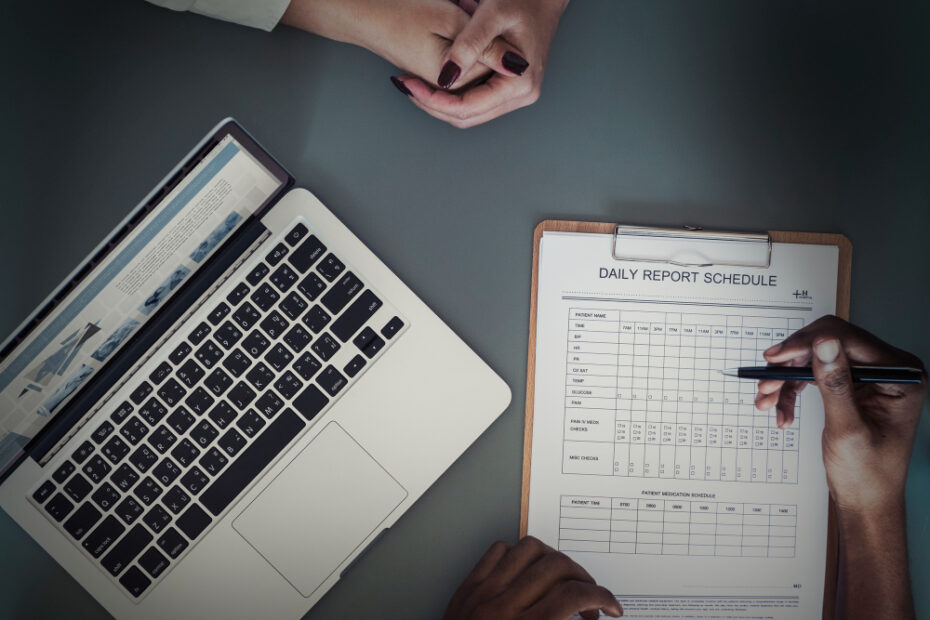 This image shows a professional setting where two individuals are working collaboratively. On the desk, there is a laptop displaying information, and a clipboard with a detailed "Daily Report Schedule" form being filled out. The form appears to be used for tracking daily activities or tasks, suggesting a focus on productivity, planning, or time management. This setup aligns well with creating or managing timesheets