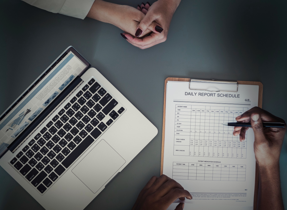 This image shows a professional setting where two individuals are working collaboratively. On the desk, there is a laptop displaying information, and a clipboard with a detailed "Daily Report Schedule" form being filled out. The form appears to be used for tracking daily activities or tasks, suggesting a focus on productivity, planning, or time management. This setup aligns well with creating or managing timesheets