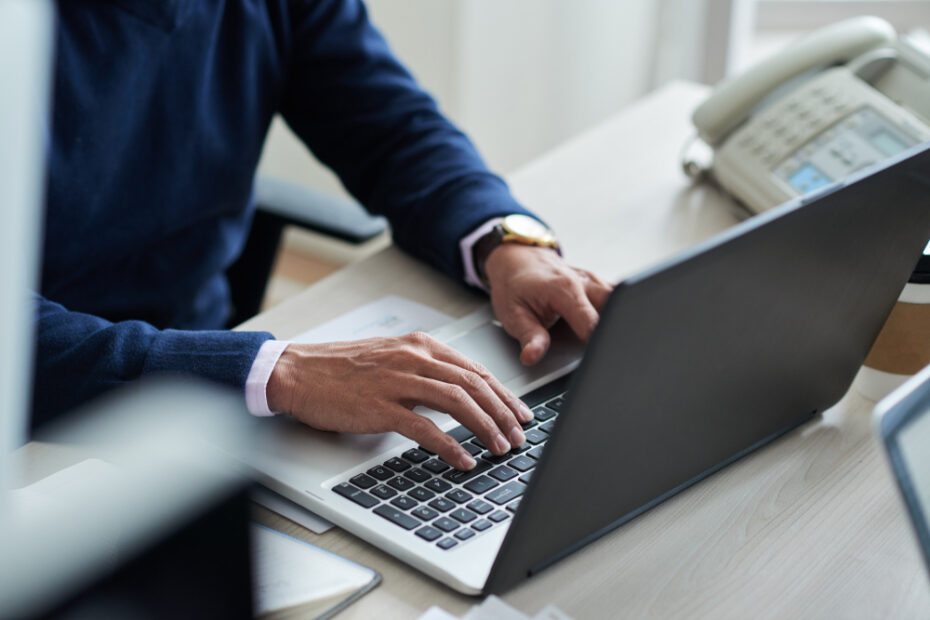 A close-up image of a person working on a laptop at their desk, with hands typing on the keyboard. The scene suggests a professional setting, with office items like a telephone and documents nearby, highlighting productivity and digital task management.