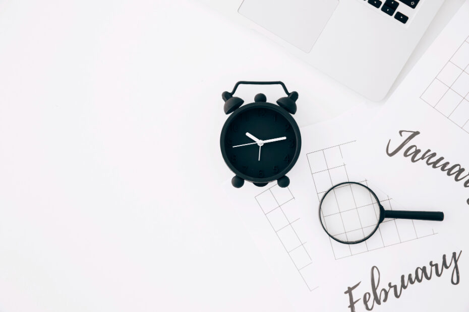 A minimalist, flat-lay setup with a black analog alarm clock, a magnifying glass, and printed calendar pages labeled "January" and "February," placed on a white surface alongside a laptop. The arrangement suggests themes of time management, scheduling, and planning, ideal for illustrating concepts related to productivity, project timelines, or PTO (Paid Time Off) tracking.