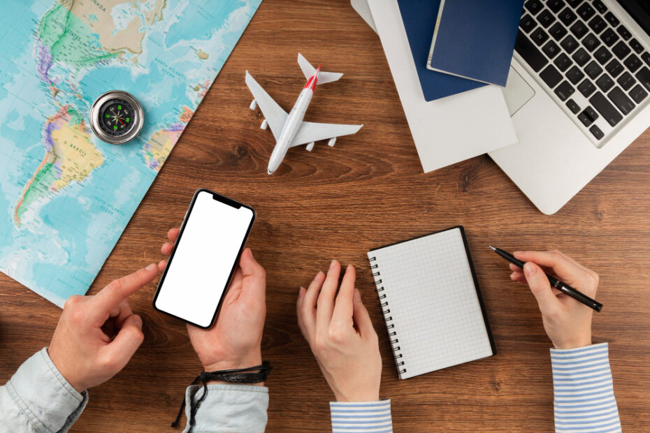 This image captures a travel planning setup on a wooden table. The scene includes a world map, a compass, a smartphone with a blank screen, a small model airplane, two passports, a laptop, and a notepad with a pen. Two people appear to be planning or discussing travel arrangements, with one person pointing at the phone, perhaps reviewing travel options or information. The composition conveys a sense of adventure, organization, and anticipation, ideal for illustrating trip preparation or itinerary planning.