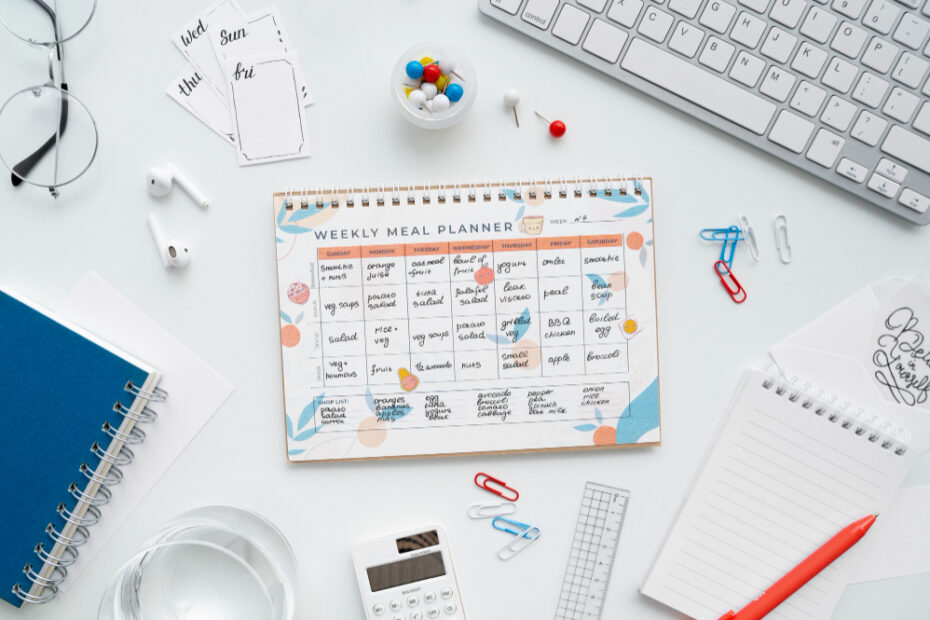 A top-down view of a workspace with a colorful weekly meal planner notebook in the center, surrounded by various office and stationery items, including a keyboard, eyeglasses, a calculator, paper clips, push pins, notebooks, and a pen. The planner displays a detailed meal schedule for each day, alongside a shopping list at the bottom.