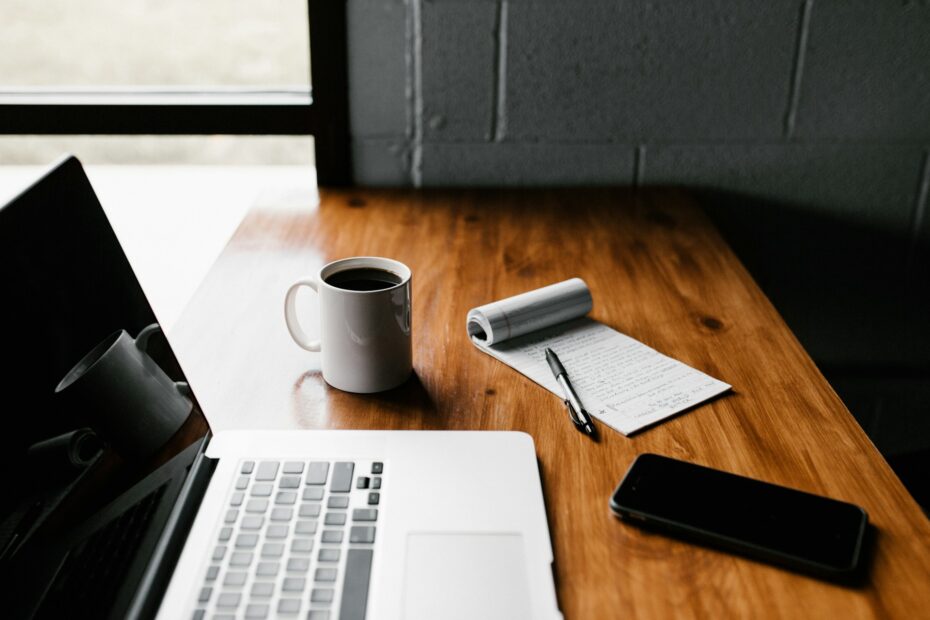 The image shows a calm, minimalist desk setup with a wooden surface, featuring an open laptop, a mug of coffee, a notebook with a pen, and a smartphone placed nearby. Natural light streams in from the adjacent window, adding a serene and focused atmosphere, ideal for work or writing. The arrangement suggests a productive workspace or study setting, with a touch of simplicity that creates an inviting environment for concentration and creativity.