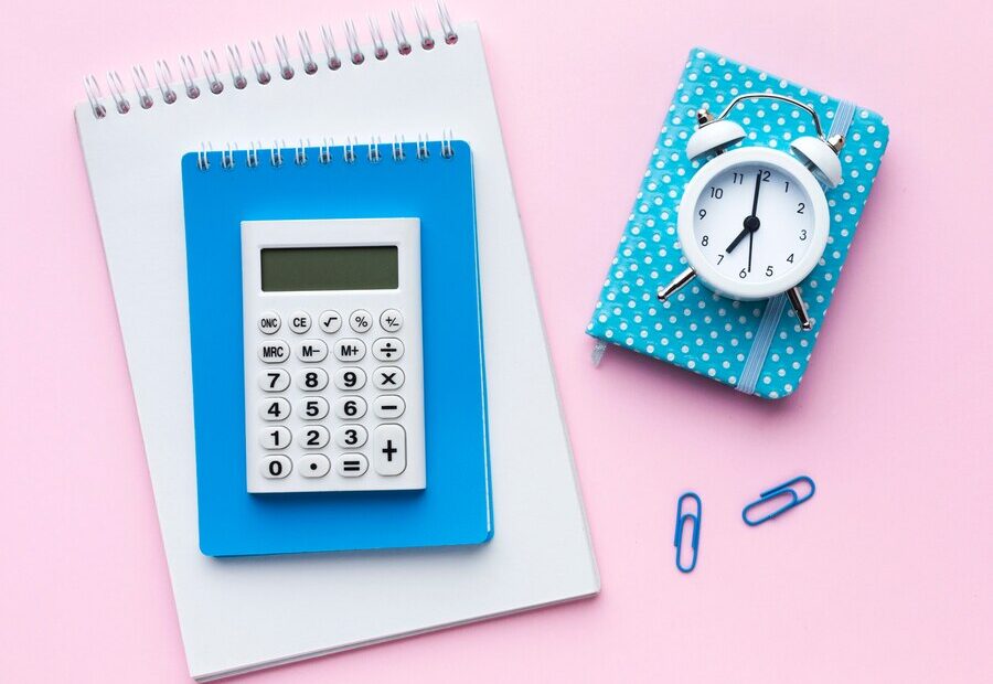 This image features a flat lay arrangement of stationery items on a pastel pink background. At the center is a white calculator resting on a blue notebook, placed on top of a white spiral-bound notepad. To the right, a small blue polka-dotted planner lies with a white alarm clock sitting on top. Blue paper clips add subtle details, creating a clean, organized, and aesthetically pleasing workspace vibe.