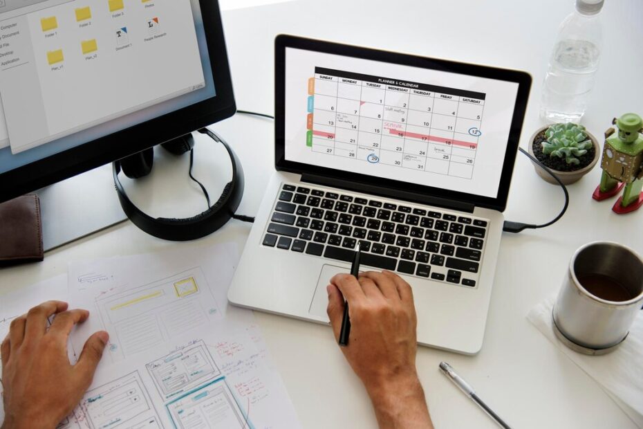 This image shows a workspace setup featuring a person working with a laptop displaying a planner or calendar on the screen, alongside a monitor showing folders and documents. The individual is holding a pen, and there are papers with sketches and notes on the desk. Additional items include a cup of coffee, a water bottle, a small potted plant, and a small robot figurine, creating an organized yet creative environment.