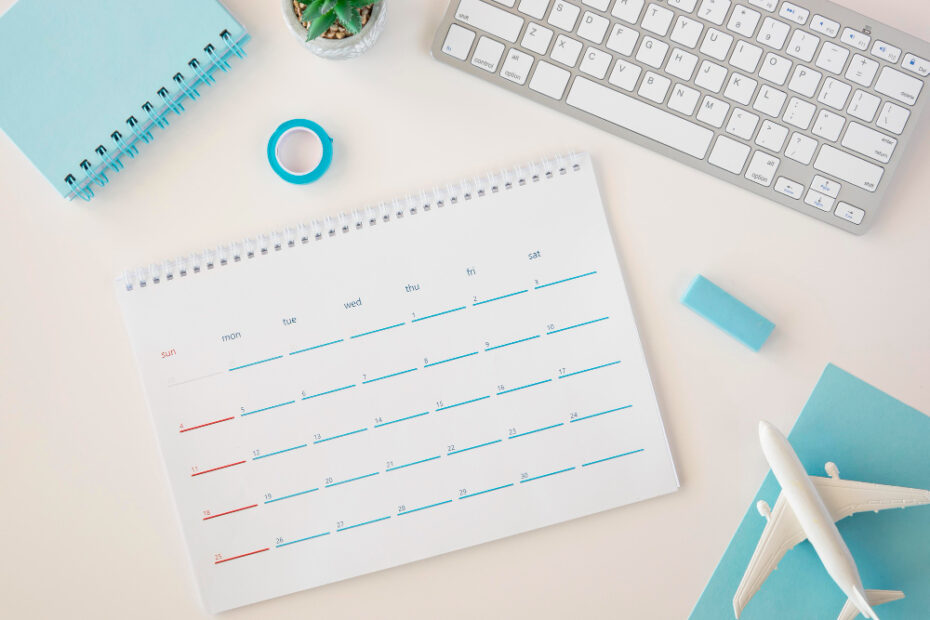 A clean, minimalist workspace featuring a desk calendar with blue and red markings, a keyboard, a small notebook, a roll of blue tape, a blue eraser, a miniature airplane model, and a small potted succulent plant. The items are arranged neatly on a white desk with a light blue theme.