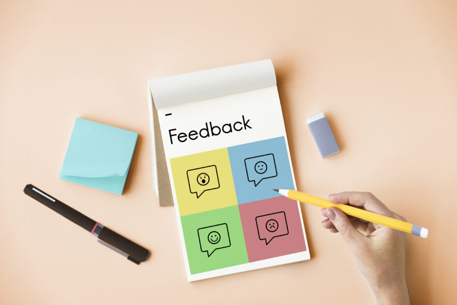 The image shows a notepad labeled "Feedback" with four colorful squares, each containing speech bubbles with various facial expressions (happy, neutral, surprised, and sad). Surrounding the notepad are a pen, sticky notes, an eraser, and a hand holding a pencil, reflecting the theme of peer review feedback and evaluation.