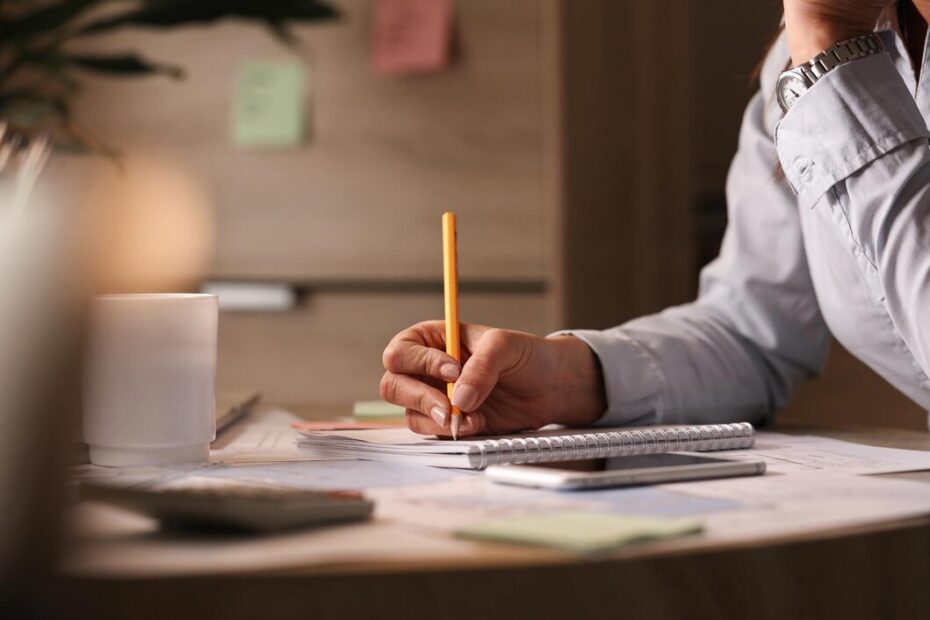 A person holding a pencil and writing in a notebook while working at a desk, surrounded by notes, a smartphone, and a coffee cup, illustrating tips for writing reports effectively.