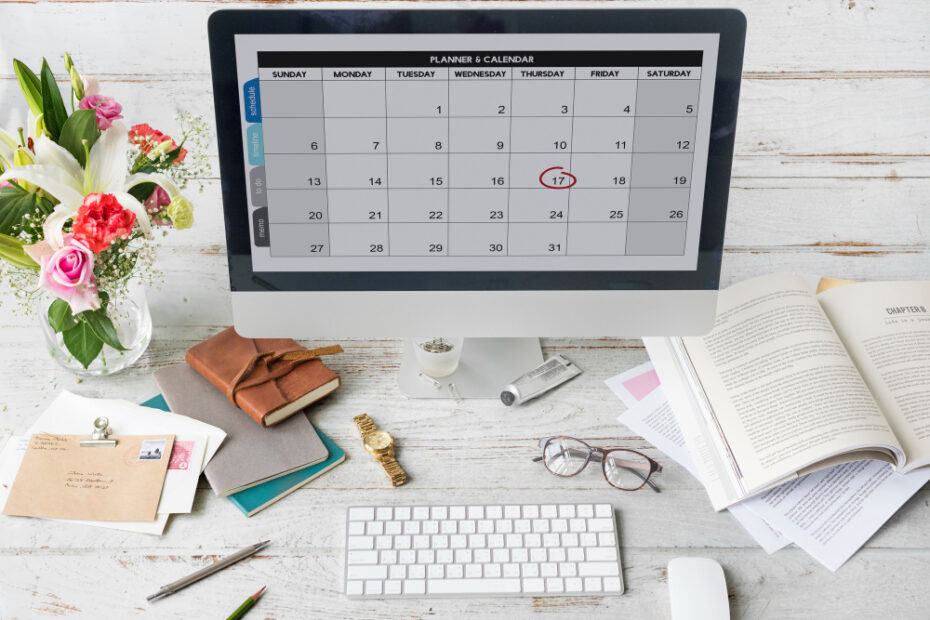 A modern workspace featuring a desktop computer displaying a digital planner and calendar, with a date circled. The desk is decorated with flowers, books, glasses, stationery, and office supplies. The setup suggests the use of a vacation tracking tool for managing schedules and planning time off efficiently.