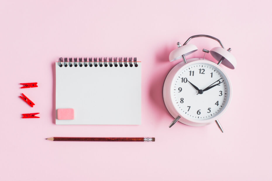 Flat lay of a leave tracker system concept with a blank notepad, pencil, red clips, eraser, and a white alarm clock on a pastel pink background.