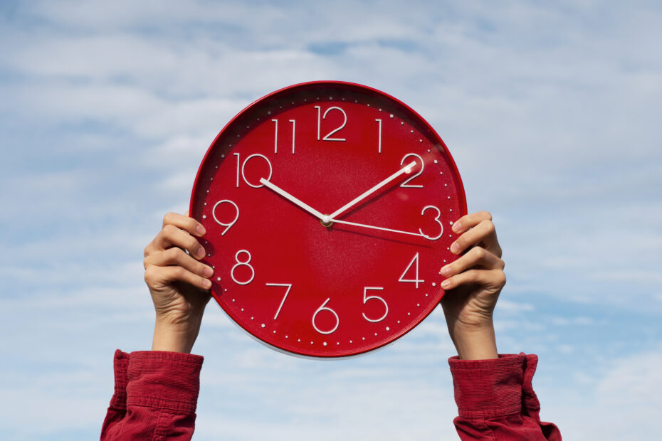 A person wearing a red jacket holds up a large red clock against a bright blue sky. The clock shows the time as 10:10. This image represents the concept of time and flexibility, relating to FTO, which stands for Flexible Time Off, a modern workplace leave policy allowing employees to take time off as needed.