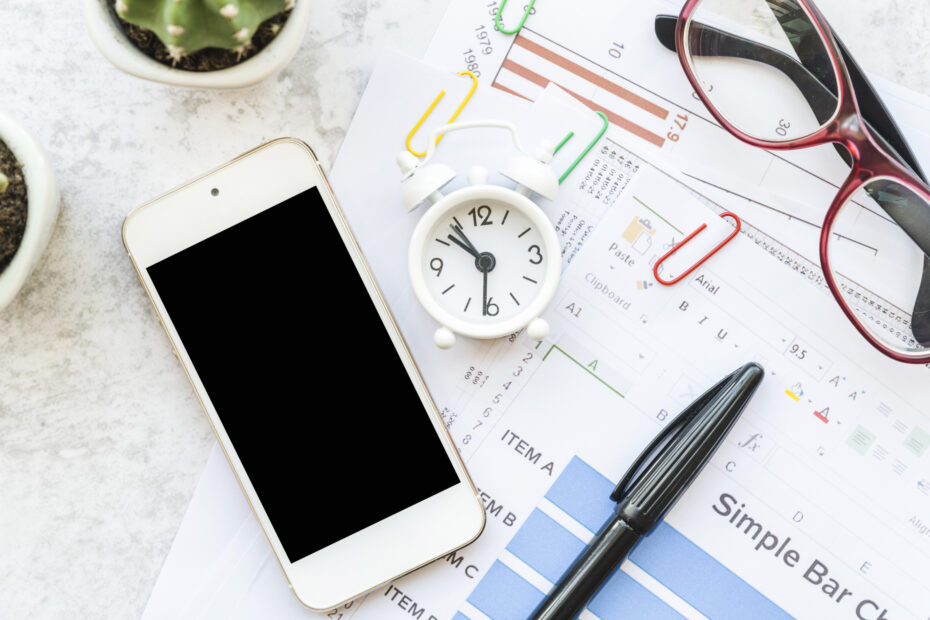 Office workspace with a smartphone, small clock, pen, and documents featuring charts and spreadsheets, representing productivity and employee time tracking app usage.