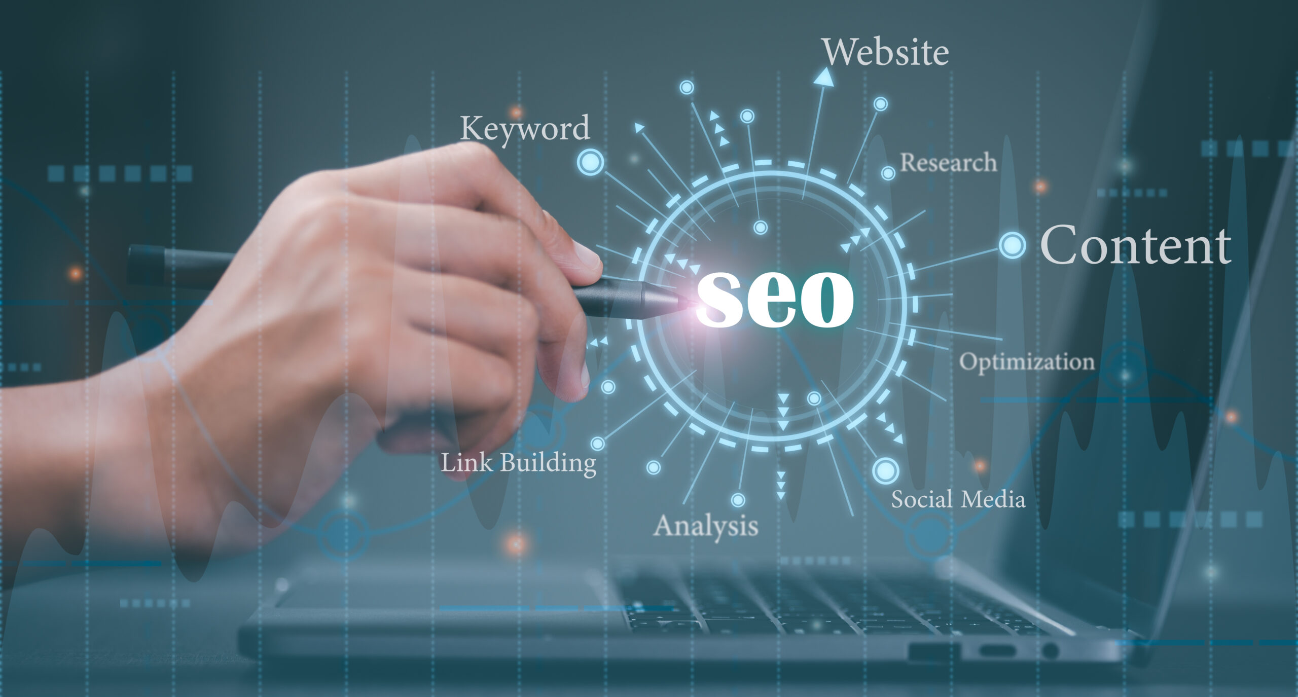 A digital representation of SEO (Search Engine Optimization) concepts, featuring a hand holding a pen pointing at the word "SEO" in a futuristic interface. Surrounding keywords such as "Website," "Content," "Keyword," "Research," "Optimization," "Social Media," "Link Building," and "Analysis" emphasize various aspects of SEO. A laptop in the background symbolizes the digital nature of SEO strategies.