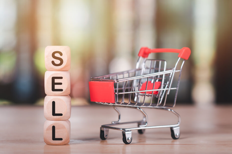 Wooden blocks spelling "SELL" stacked vertically next to a small shopping cart with red accents, symbolizing commerce and transactions. The image represents selling to local businesses and the concept of retail, sales, and business growth.